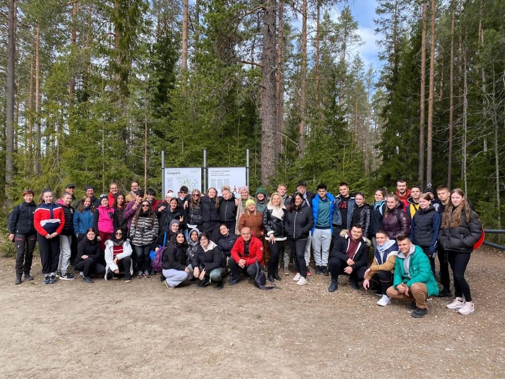 Ryhmä opiskleijoita ja opettajia kuvattuna metsäisessä maisemassa Geoparkin kyltin vieressä.