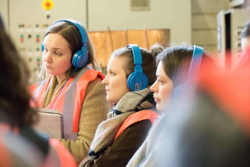 Kolme henkilöä kuuntelee kuulokkeiden kautta tarkkaavaisesti esitystä panimovierailun yhteydessä.