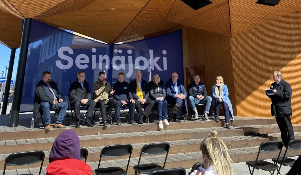 Yhdeksän panelistia istumassa Seinäjoen toripaviljongissa ja juontaja seisomassa heidän oikealla puolellaan.