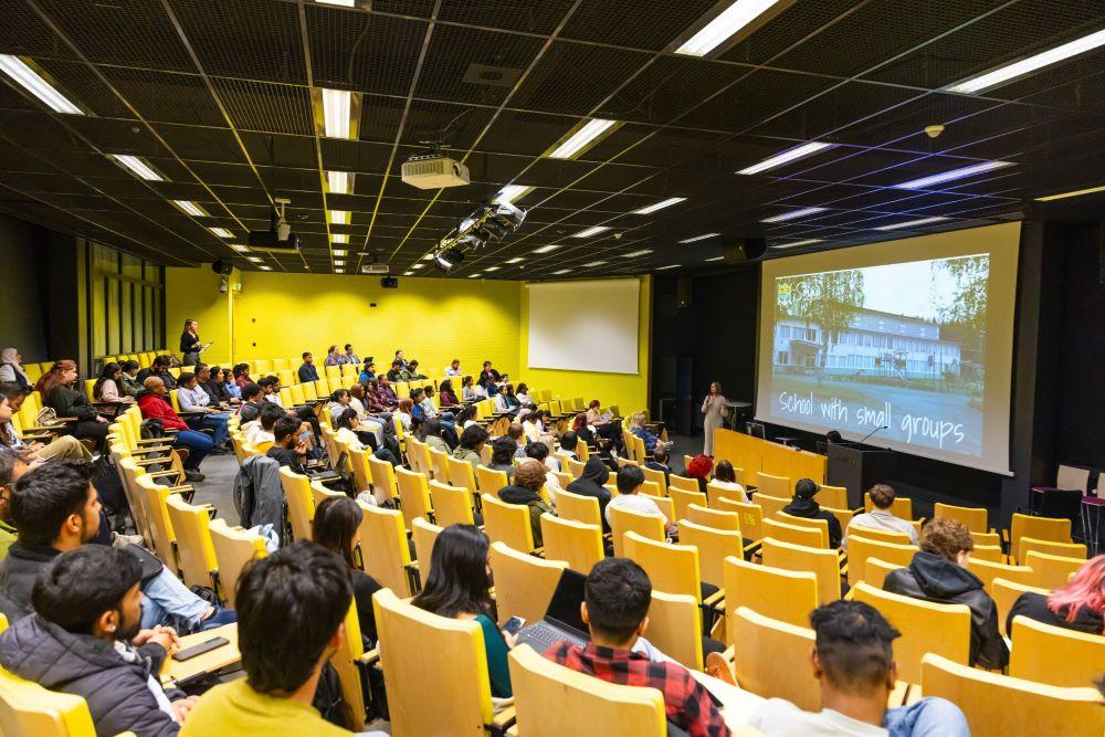 Ihmisiä auditoriossa kuuntelemassa esitystä. People in the auditorium listening to a presentation.