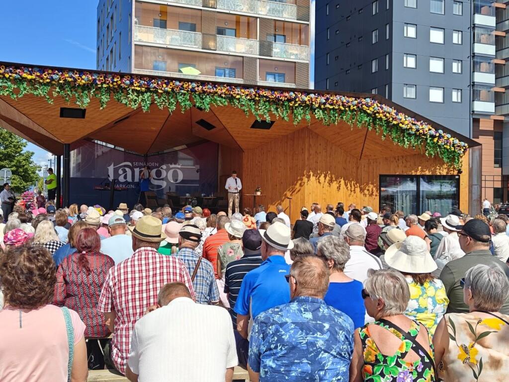 Ihmisiä istumassa penkillä ja kuuntelemassa puhujaa lavalla.